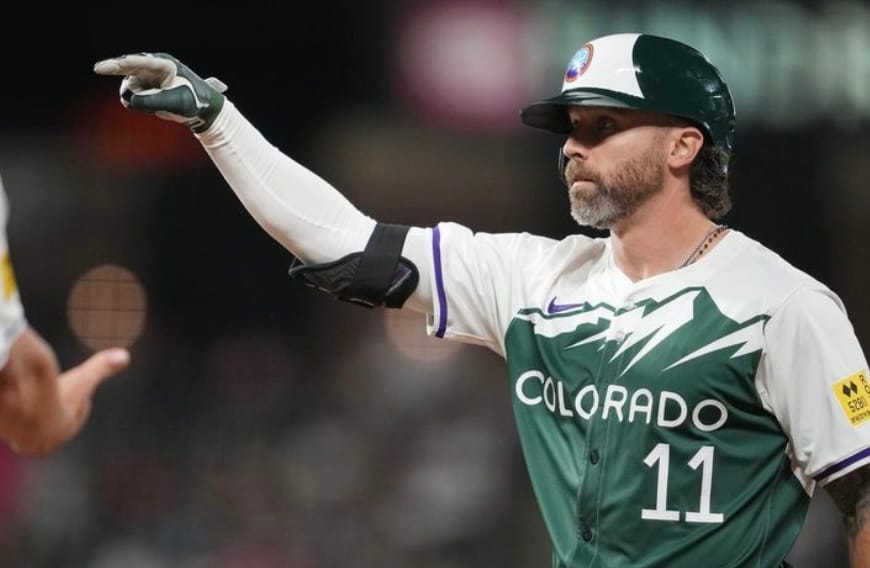 Cave stands on base, pointing with his right hand. He’s wearing the Rockies City Connects.