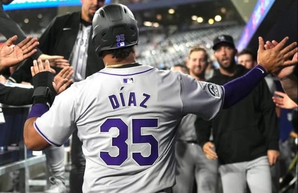 Elias Díaz high fives his teammates in the dugout after scoring a run. He’s wearing a gray uniform.